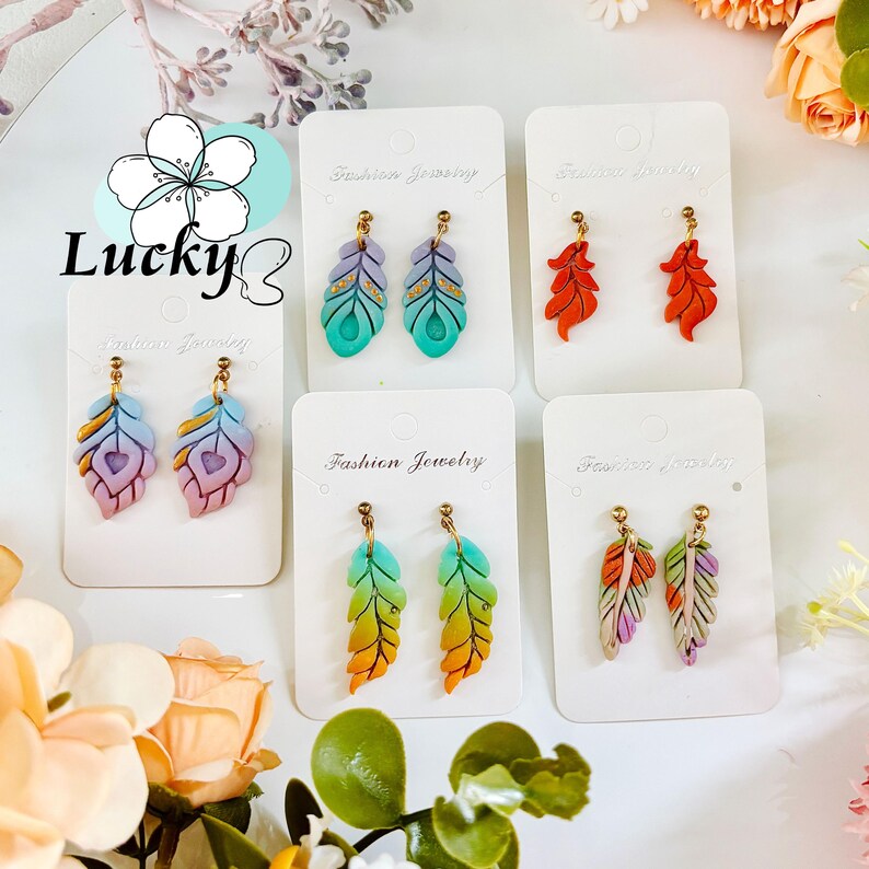 May include: A selection of feather-shaped earrings in shades of blue, orange, purple, and green. Each pair is presented on a white card marked "Fashion Jewelry" with gold-tone earring hooks. The word "Lucky" is visible in the image.