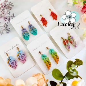 May include: Feather-shaped earrings in shades of orange, green, purple, and blue, presented on white cards. Each earring features a gold-coloured post. The cards are printed with the text "Fashion Jewelry". The word "Lucky" is in the image.