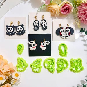 May include: A selection of Halloween-themed earrings and cookie cutters. The earrings showcase spooky designs such as ghosts, bats, and skull characters. The cookie cutters are bright green and shaped like ghosts and other Halloween figures.