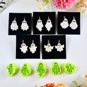 May include: Handmade ghost earrings on black cards. The white earrings have gold-coloured hooks. Below are green ghost-shaped cookie cutters. The earrings and cutters are likely for Halloween, a spooky holiday.