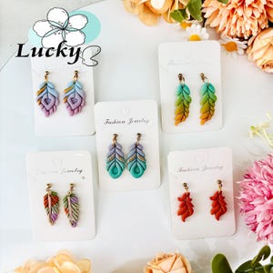May include: A selection of feather-shaped earrings in various colours, including blue, green, orange, and red, with gold-coloured details. The earrings are displayed on white cards. The background features floral arrangements.