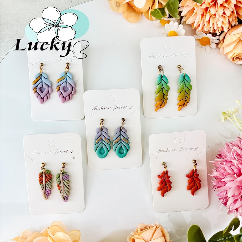 May include: A selection of feather-shaped earrings in various colours, including blue, green, orange, and red, with gold-coloured details. The earrings are displayed on white cards. The background features floral arrangements.