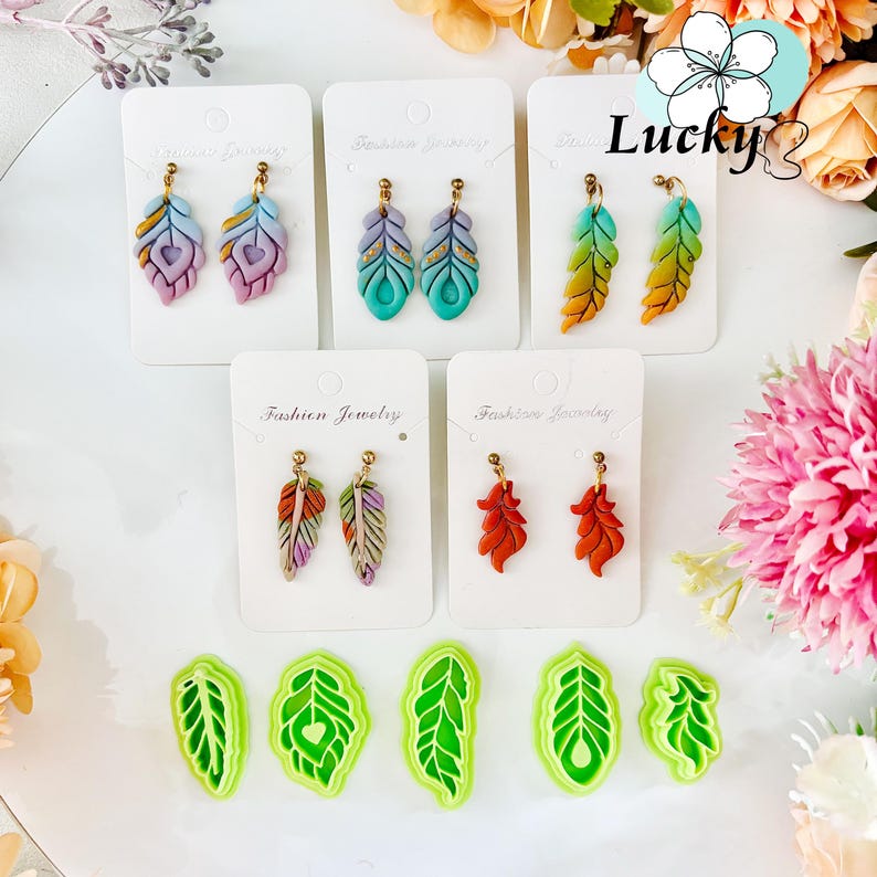 May include: A selection of handmade feather earrings in shades of purple, teal, orange, and red, presented on white cards. The earrings feature gold-coloured posts. Green leaf-shaped cutters are at the bottom. The word "Lucky" is visible.