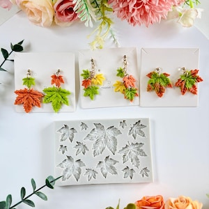 May include: Three pairs of leaf earrings in shades of orange, green, and yellow are displayed on white cards. A silicone mould with leaf impressions is below. The earrings have gold-coloured hooks. The background includes flowers.