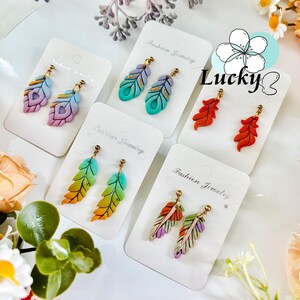 May include: A selection of feather-shaped earrings in shades of purple, blue, green, orange, and red. Each pair is presented on a white card labelled "Fashion Jewelry" with gold-coloured earring posts.