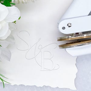 May include: A white metal embosser tool with brass plates positioned on a piece of white paper. The paper features a debossed monogram of the letters S and B in a cursive script. White artificial flowers are visible on the left side of the frame.