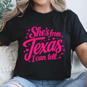 K&ouml;nnte beinhalten: Schwarzes T-Shirt mit dem Text "She's from Texas I can tell" in leuchtendem Pink. Der Text ist in einer Schreibschrift mit Herzen und Sternen gehalten. Das T-Shirt hat einen l&auml;ssigen Stil.