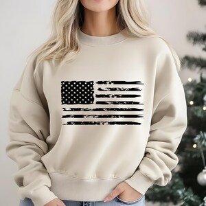 May include: A beige sweatshirt featuring a distressed black and white American flag design. The flag graphic includes stars and stripes, giving a vintage or worn look. The sweatshirt is a neutral color, suitable for casual wear.