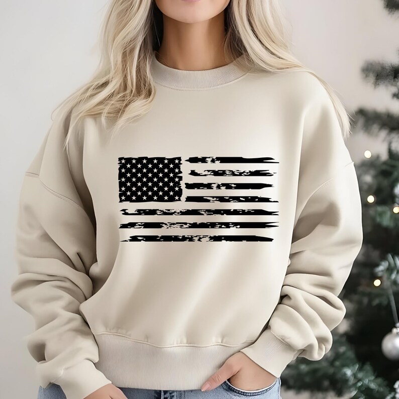May include: A beige sweatshirt featuring a distressed black and white American flag design. The flag graphic includes stars and stripes, giving a vintage or worn look. The sweatshirt is a neutral color, suitable for casual wear.