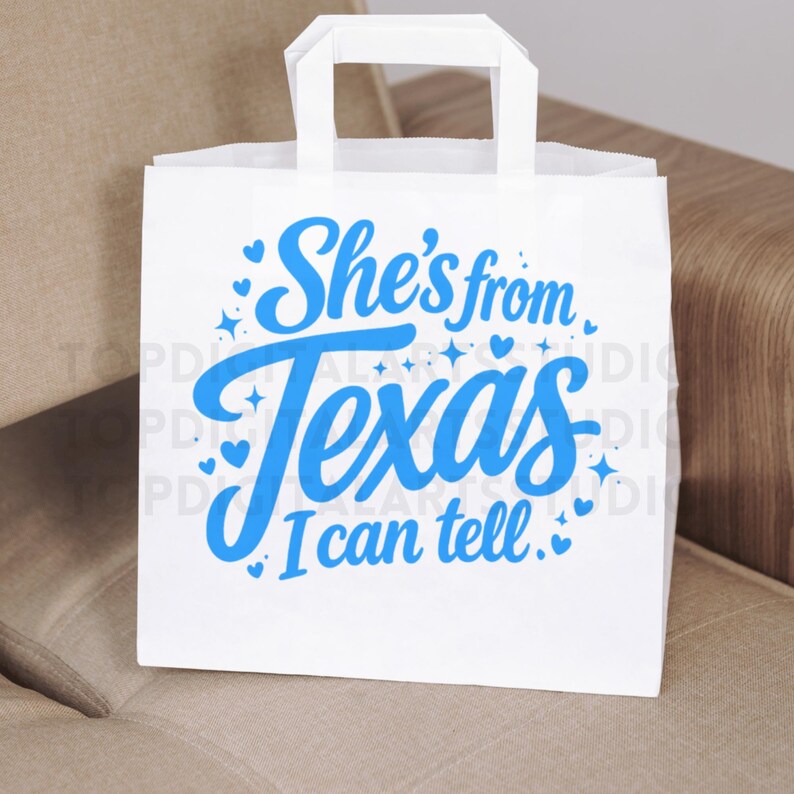 K&ouml;nnte beinhalten: Wei&szlig;e Papiert&uuml;te mit rechteckigem Griff. Auf der T&uuml;te steht in blauer Schrift "She's from Texas I can tell" sowie dekorative Herzen und Sterne. Die T&uuml;te steht auf einer beigen Oberfl&auml;che.