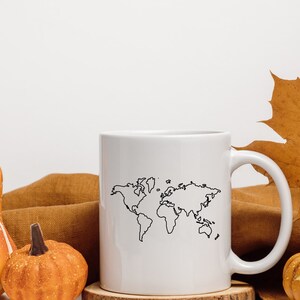 K&ouml;nnte beinhalten: Eine wei&szlig;e Keramiktasse mit einer schwarzen Umrandung einer Weltkarte. Die Tasse steht auf einer braunen Oberfl&auml;che mit zwei kleinen orangefarbenen K&uuml;rbissen und einem gro&szlig;en orangefarbenen K&uuml;rbis mit wei&szlig;en Flecken.