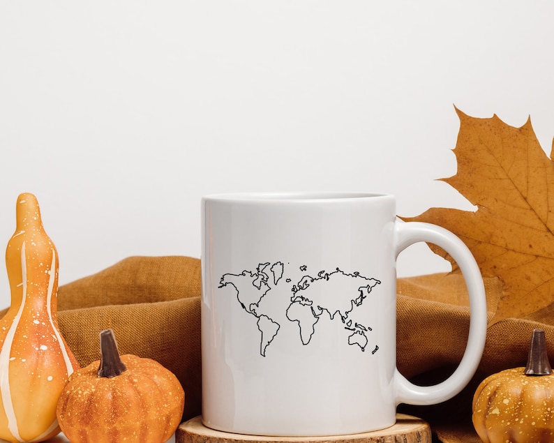 K&ouml;nnte beinhalten: Eine wei&szlig;e Keramiktasse mit einer schwarzen Umrandung einer Weltkarte. Die Tasse steht auf einer braunen Oberfl&auml;che mit zwei kleinen orangefarbenen K&uuml;rbissen und einem gro&szlig;en orangefarbenen K&uuml;rbis mit wei&szlig;en Flecken.