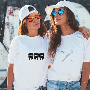 May include: Two friends wearing white t-shirts with dental-themed graphics. One shirt features a graphic of three teeth with braces, while the other shirt features a graphic of crossed dental tools.