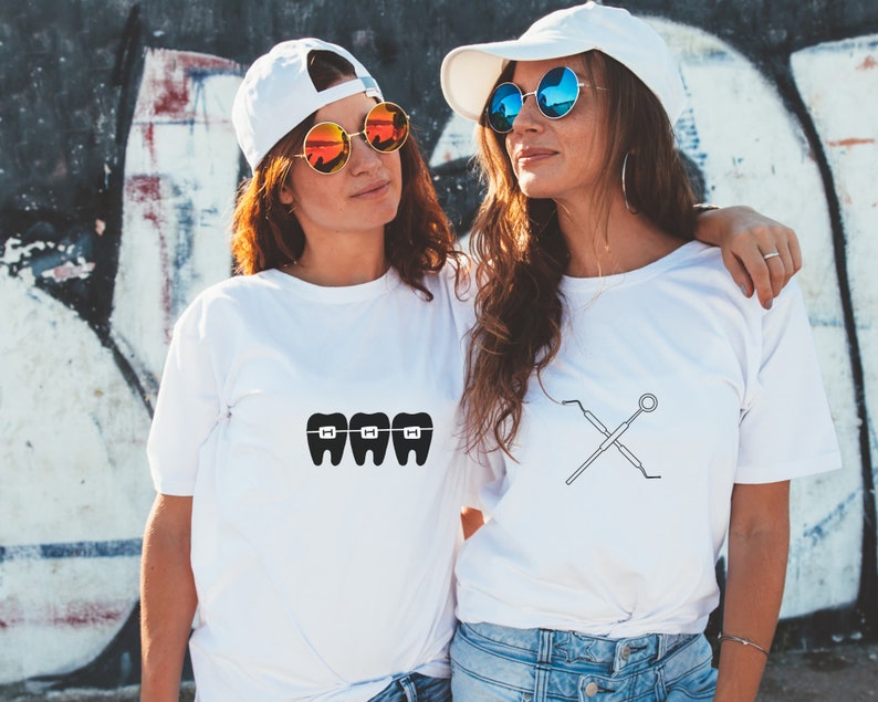 May include: Two friends wearing white t-shirts with dental-themed graphics. One shirt features a graphic of three teeth with braces, while the other shirt features a graphic of crossed dental tools.