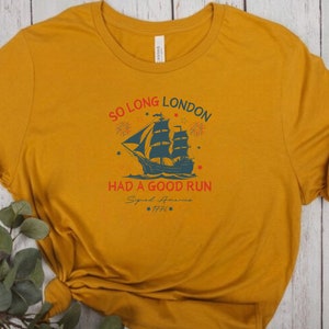 May include: A mustard yellow t-shirt with a vintage-style graphic of a ship sailing away from a city skyline. The text reads "So Long London Had A Good Run Signed America 1776".