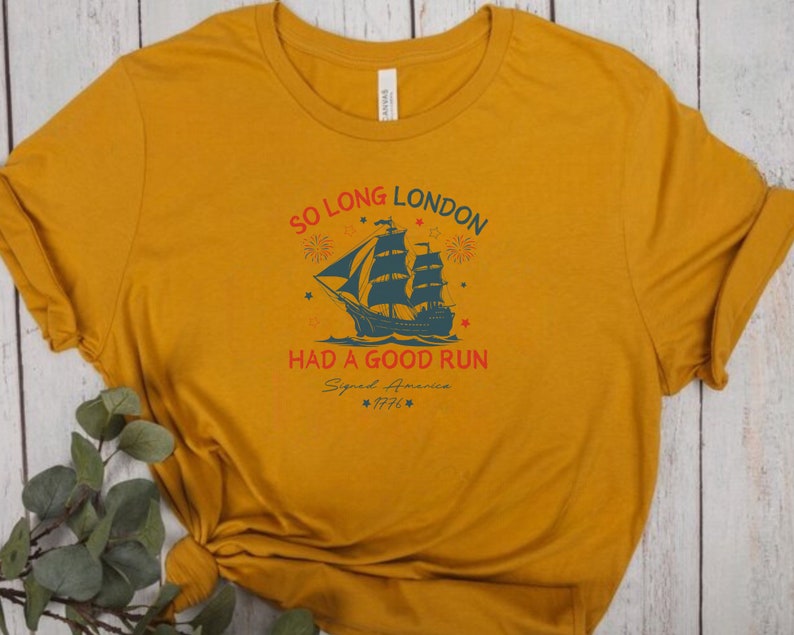 May include: A mustard yellow t-shirt with a vintage-style graphic of a ship sailing away from a city skyline. The text reads "So Long London Had A Good Run Signed America 1776".