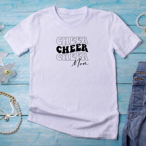 May include: White t-shirt with black text that reads "Cheer Cheer Cheer Mom" in a retro font.
