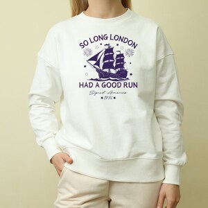 May include: White sweatshirt with a navy blue graphic of a ship sailing away from a city skyline with fireworks. The text "So Long London Had A Good Run" is printed on the shirt with the year "1776" below.