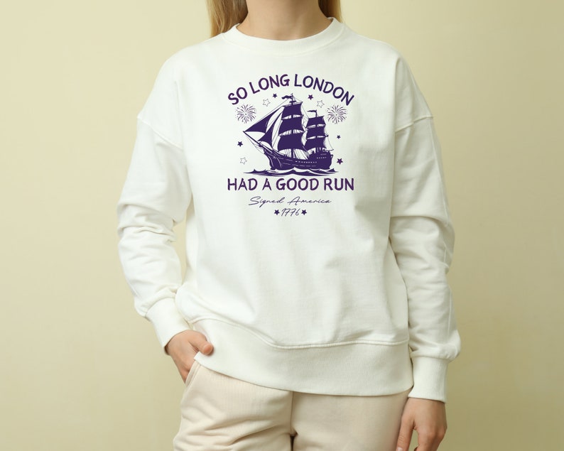 May include: White sweatshirt with a navy blue graphic of a ship sailing away from a city skyline with fireworks. The text "So Long London Had A Good Run" is printed on the shirt with the year "1776" below.