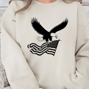 May include: A cream-colored sweatshirt featuring a black graphic of an eagle with outstretched wings, clutching an American flag. The eagle is detailed with white accents, and the flag has a star field and stripes.