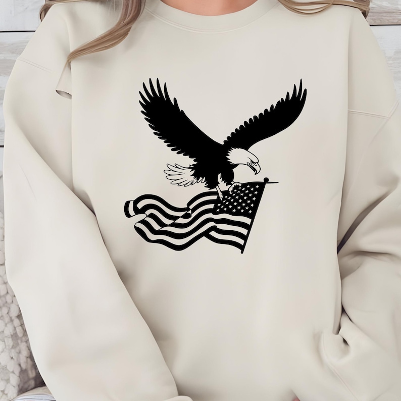 May include: A cream-colored sweatshirt featuring a black graphic of an eagle with outstretched wings, clutching an American flag. The eagle is detailed with white accents, and the flag has a star field and stripes.