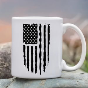 May include: White ceramic mug featuring a distressed black graphic of the American flag. The flag design includes stars and stripes, with a worn, vintage aesthetic. The mug has a curved handle and is suitable for hot or cold beverages.