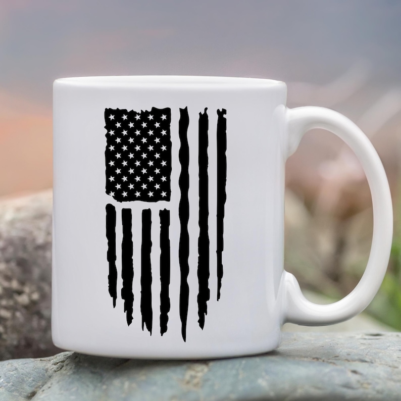 May include: White ceramic mug featuring a distressed black graphic of the American flag. The flag design includes stars and stripes, with a worn, vintage aesthetic. The mug has a curved handle and is suitable for hot or cold beverages.