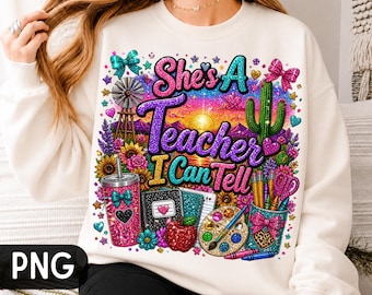 Puede incluir: Una sudadera color crema con un gráfico brillante que incluye el texto "She's A Teacher I Can Tell". El diseño muestra un molino de viento, un cactus, girasoles, un vaso con purpurina, un cuaderno, una manzana, una paleta y lápices.