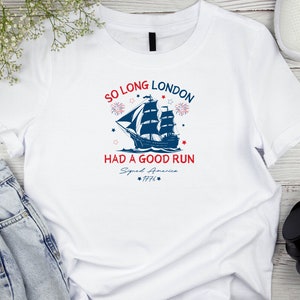 May include: White t-shirt with a blue and red graphic of a ship sailing away from a city skyline. The text "So Long London Had A Good Run Signed America 1776" is printed on the shirt.