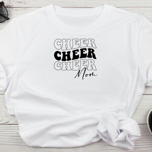 May include: White t-shirt with the text "Cheer Cheer Cheer Mom" in black retro font.