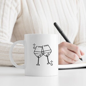 May include: White ceramic mug with a black line drawing of two wine glasses clinking together. The mug is on a white surface.