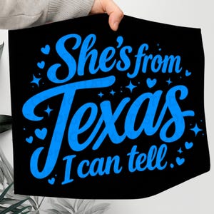 K&ouml;nnte beinhalten: Schwarzes Textil mit dem Text "She's from Texas, I can tell" in leuchtend blauer Schrift. Das Design umfasst Herz- und Sternakzente. Der Artikel wird von einer Person gehalten.