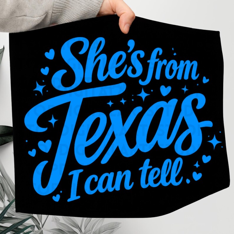 K&ouml;nnte beinhalten: Schwarzes Textil mit dem Text "She's from Texas, I can tell" in leuchtend blauer Schrift. Das Design umfasst Herz- und Sternakzente. Der Artikel wird von einer Person gehalten.