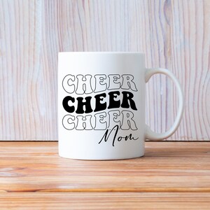 May include: White ceramic mug with black text that reads "Cheer Cheer Cheer Mom" in a retro font.