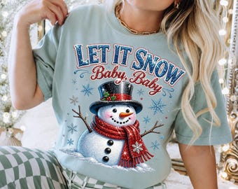 Puede incluir: Una camiseta azul claro con el gráfico de un muñeco de nieve con sombrero de copa negro y bufanda roja. El texto "LET IT SNOW Baby, Baby" está impreso encima en letras brillantes azules y rojas, rodeado de pequeños copos de nieve.