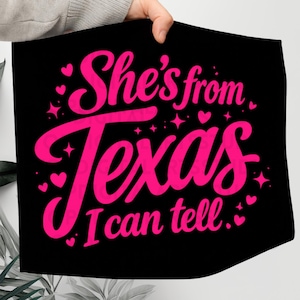 K&ouml;nnte beinhalten: Schwarzes Textil mit dem Text "She's from Texas I can tell" in leuchtendem Pink, umgeben von Herzen und Sternen. Das Design ist in einer verspielten, kursiven Schriftart gehalten.