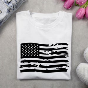 May include: White t-shirt featuring a distressed black American flag design. The flag graphic includes stars and stripes, giving a vintage or worn look. The shirt is folded and placed on a gray surface, with a pair of jeans and sneakers visible.