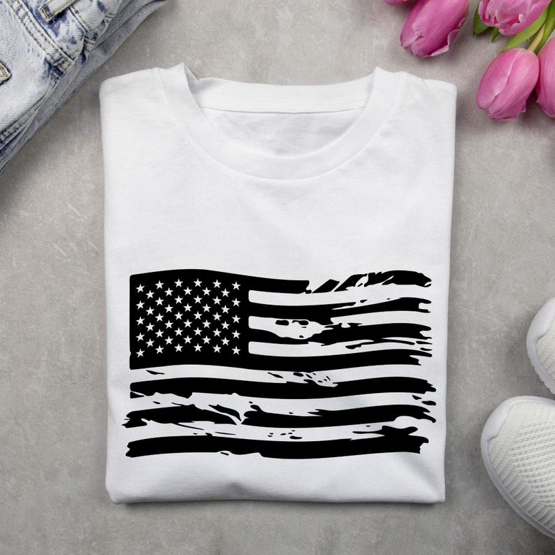 May include: White t-shirt featuring a distressed black American flag design. The flag graphic includes stars and stripes, giving a vintage or worn look. The shirt is folded and placed on a gray surface, with a pair of jeans and sneakers visible.