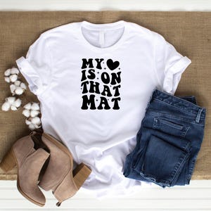 May include: White t-shirt with black text that reads "My &hearts; is on that mat". The shirt is paired with blue jeans and brown ankle boots.