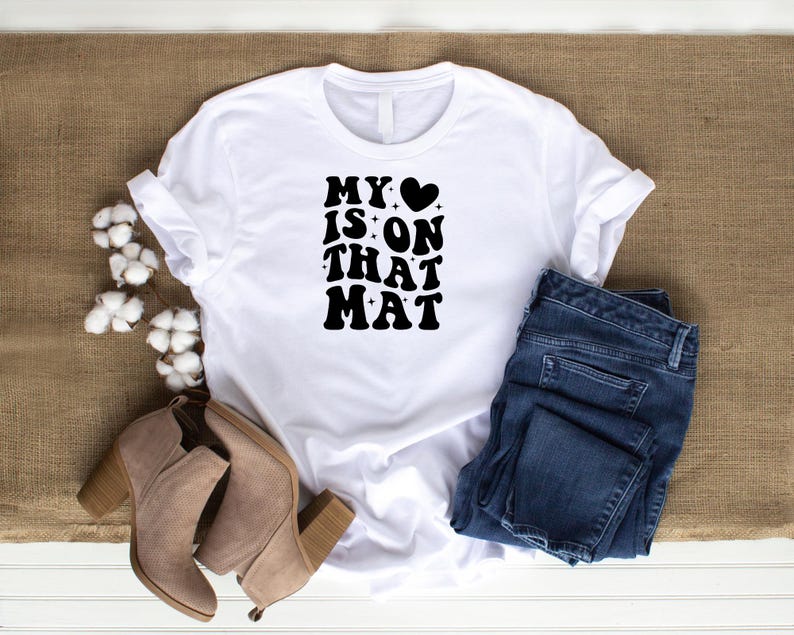 May include: White t-shirt with black text that reads "My &hearts; is on that mat". The shirt is paired with blue jeans and brown ankle boots.