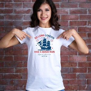 May include: White t-shirt with a blue and red graphic of a ship sailing away from a city skyline. The text on the shirt reads "So Long London Had a Good Run Squad America 1776".