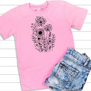 May include: A pink t-shirt with a black graphic of a bouquet of flowers with hearts. The flowers are stylized and include a sunflower, dandelions, and other blooms.