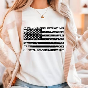 May include: White sweatshirt featuring a distressed black and white American flag design. The flag graphic includes stars and stripes, giving a vintage or worn look. The sweatshirt is a casual top.