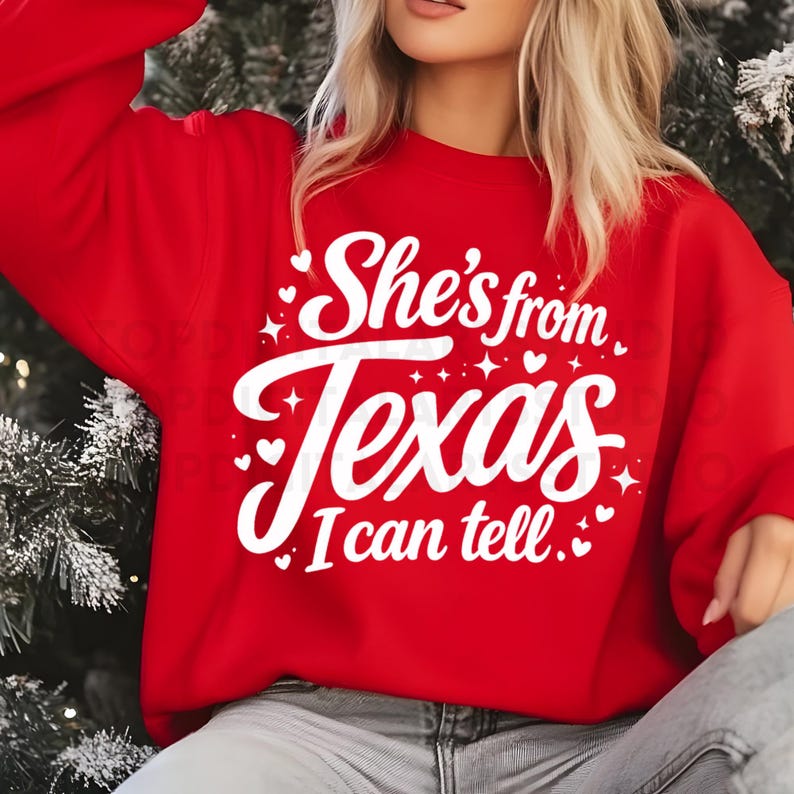 K&ouml;nnte beinhalten: Ein rotes Sweatshirt mit dem wei&szlig;en Text "She's from Texas I can tell". Das Design umfasst Herzen und Sterne. Das Sweatshirt ist ein l&auml;ssiger Stil.