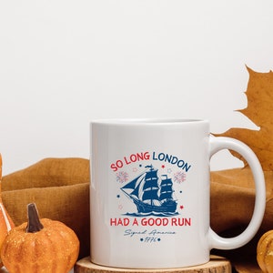 May include: White ceramic coffee mug with a blue and red graphic of a ship sailing away from fireworks. The text reads "So Long London Had a Good Run Signed America 1776".