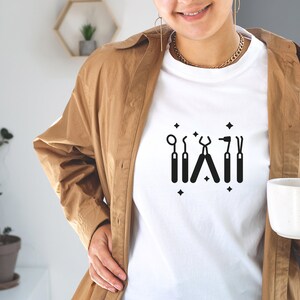 May include: White t-shirt with a black graphic of dental tools, including a mirror, a probe, pliers, a drill, and a scaler. The graphic is surrounded by small black stars.