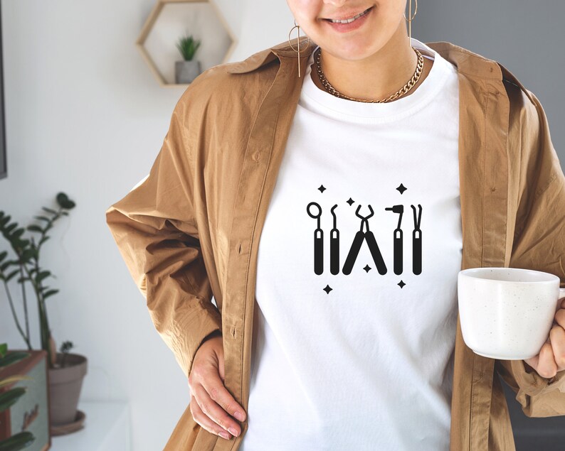 May include: White t-shirt with a black graphic of dental tools, including a mirror, a probe, pliers, a drill, and a scaler. The graphic is surrounded by small black stars.
