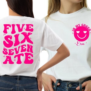 May include: Two white t-shirts with pink text and graphics. The front of one shirt says "In My Cheer Era" with a pink smiley face with hearts for eyes. The back of the other shirt says "Five Six Seven Ate" in a retro font.