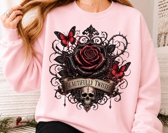 Puede incluir: Una sudadera de cuello redondo rosa claro con un gráfico central de una rosa roja oscura, un cráneo humano y dos mariposas rojas rodeadas de enredaderas negras con espinas. Una cinta contiene el texto "BEAUTIFULLY TWISTED" en color negro.