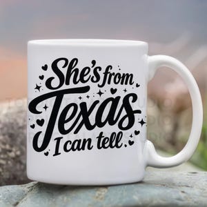 K&ouml;nnte beinhalten: Wei&szlig;e Keramiktasse mit schwarzem Schriftzug "She's from Texas, I can tell", verziert mit Herzen und Sternen. Die Tasse hat einen gebogenen Henkel und steht vor einem verschwommenen Hintergrund im Freien.